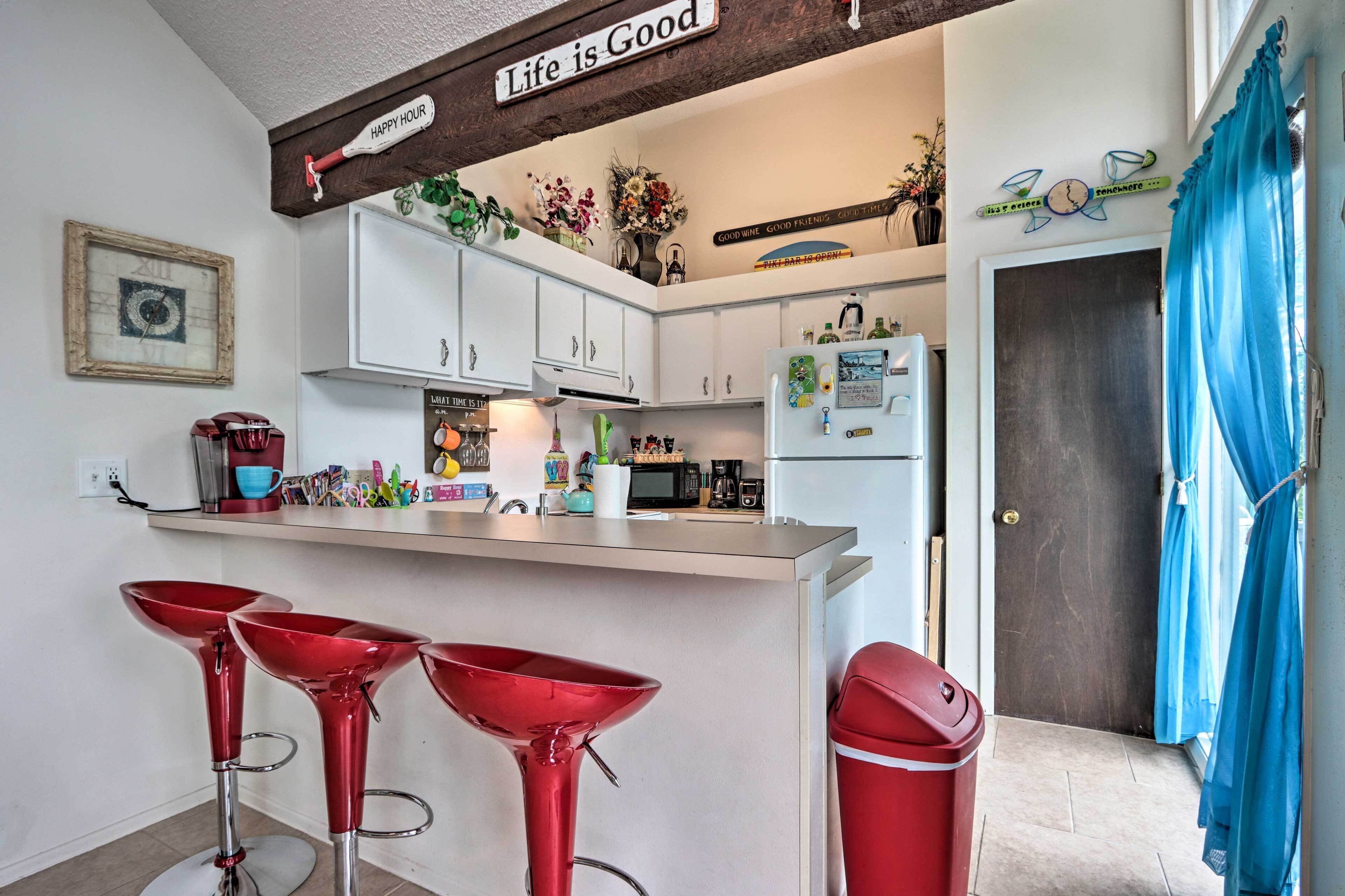 A kitchen with white cabinets, a stainless steel refrigerator, a coffee maker, and a microwave. There are red bar stools and a trash can.