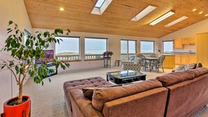 A spacious living area with a large sofa, a coffee table, and a potted plant. There are multiple windows providing views of the outdoors. - Sanderling Sea Cottages, Unit A: Pet Friendly! (Waldport)