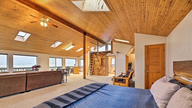 A spacious room with a large bed, a wooden ceiling, skylights, a balcony, and a view of the ocean. - Sanderling Sea Cottages, Unit A: Pet Friendly! (Waldport)