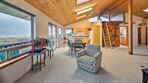A spacious living area with a view of the beach, a wooden ceiling, and a kitchen in the background. - Sanderling Sea Cottages, Unit A: Pet Friendly! (Waldport)