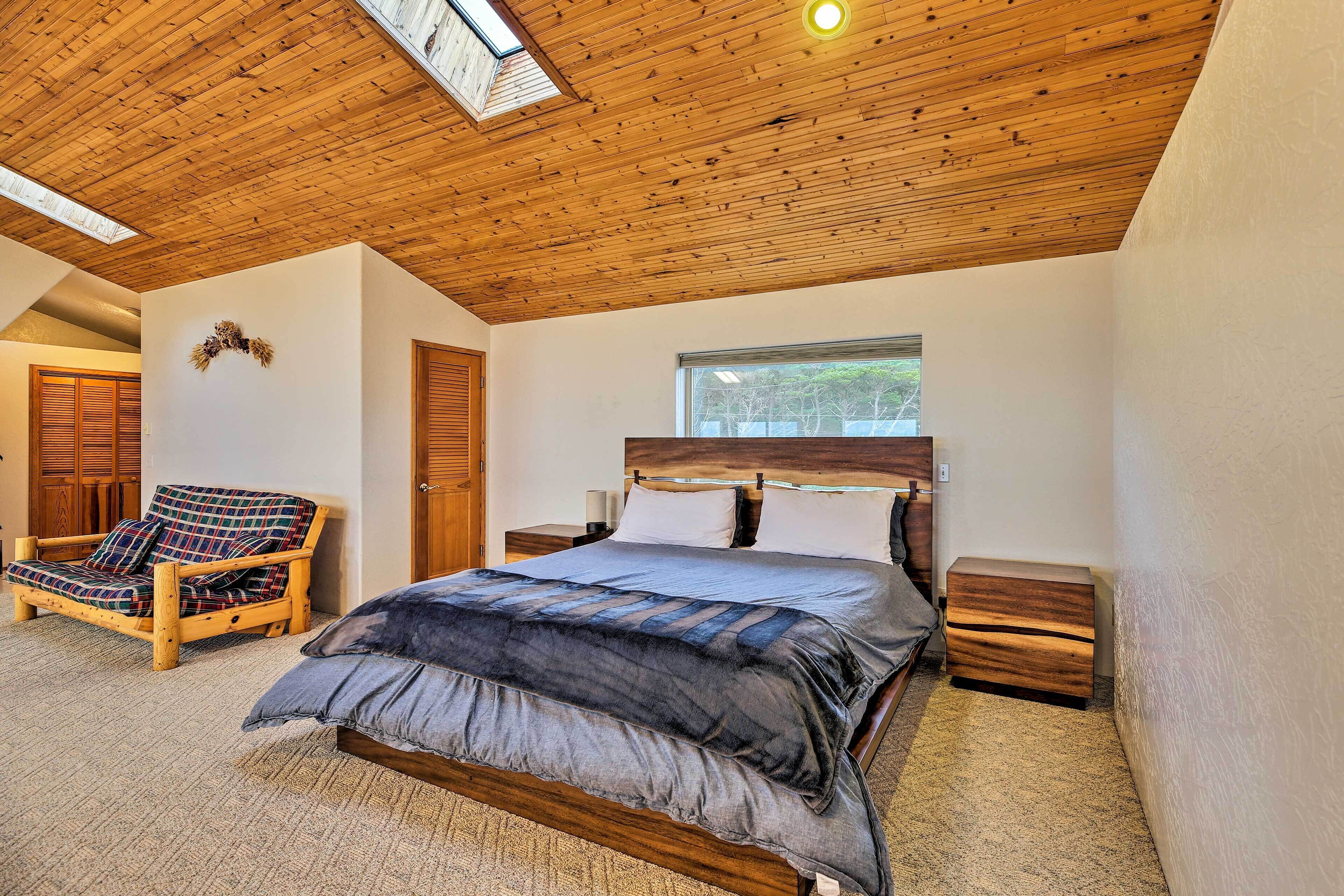 A bedroom with a wooden ceiling, a bed with a grey and blue duvet, a wooden chair, and a window with a view of trees.