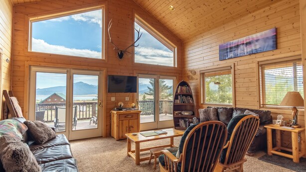 A spacious living room with a wooden ceiling, large windows, a fireplace, a television, a bookshelf, and a leather couch. - Cozy Henrys Lake Escape - 20 Miles to Yellowstone! (Island Park)