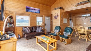 A cozy log cabin interior with a wooden ceiling, a television, a bookshelf, a couch, a coffee table, and a dining area with wooden chairs and a table. - Cozy Henrys Lake Escape - 20 Miles to Yellowstone! (Island Park)