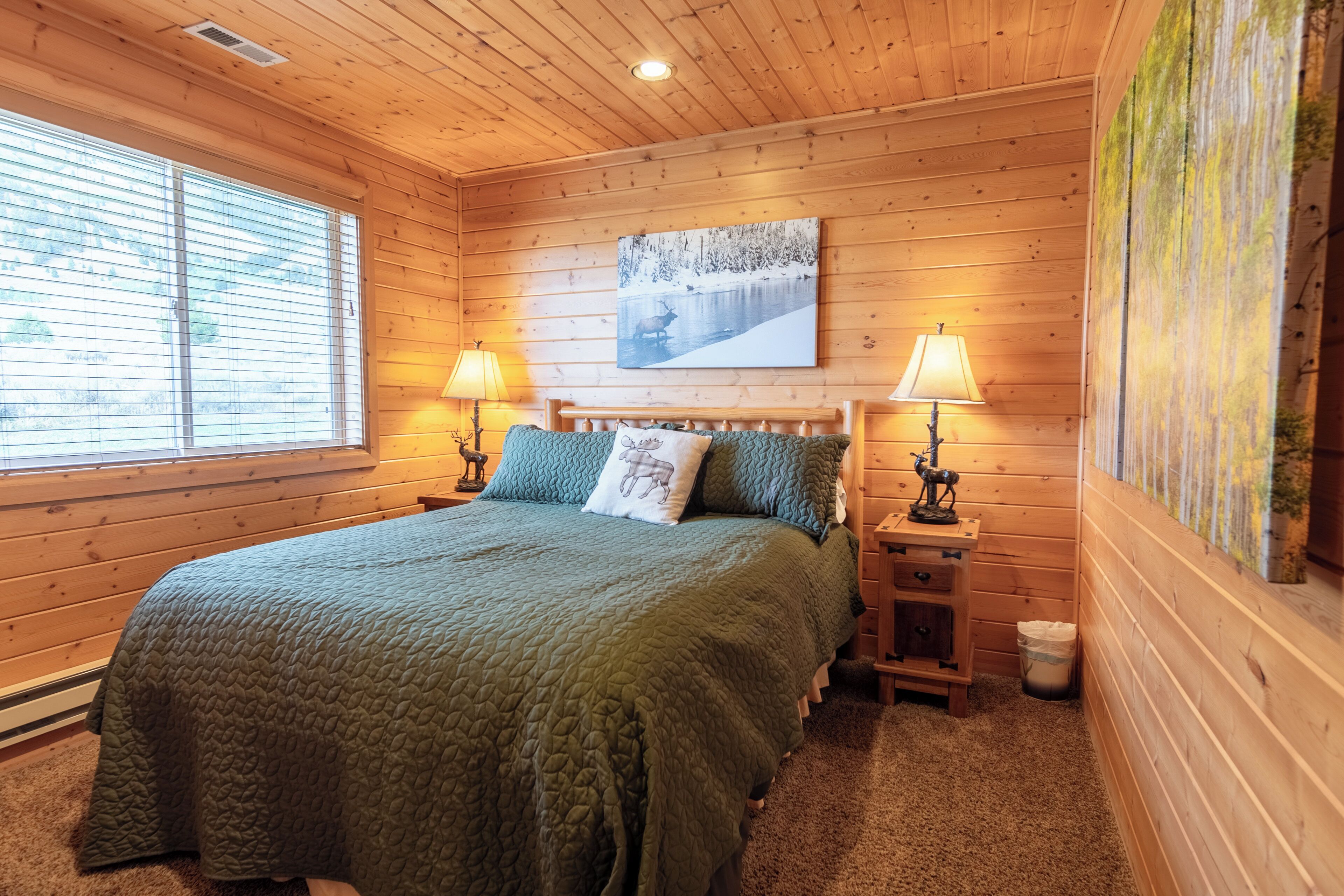 Ein Schlafzimmer mit Holzwandverkleidung, einem großen Bett, zwei Nachttischlampen, einem Bild einer verschneiten Landschaft und einem Fenster mit Jalousien.