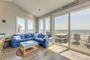 A modern living room with a large sectional sofa, a wooden bench, and a view of the beach through large sliding glass doors. - Beachfront Oasis w/ 2 Large Decks, BBQ & Views! (Holly Ridge)