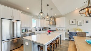 A modern kitchen with stainless steel appliances, a central island with stools, pendant lighting, and a dining area with a wooden table. - Beachfront Oasis w/ 2 Large Decks, BBQ & Views! (Holly Ridge)