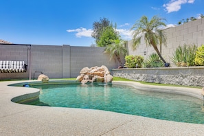 A swimming pool with a rock feature, surrounded by a low stone wall and a grassy area with palm trees. - Private Pool & Patio: Home in Surprise! (Surprise)