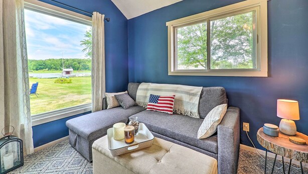 A cozy room with a blue wall, a gray sofa, a small table with a lamp, and a window offering a view of a green lawn and trees. - Cottage on Tubbs Lake w/ Fire Pit Near Skiing! (Mecosta)