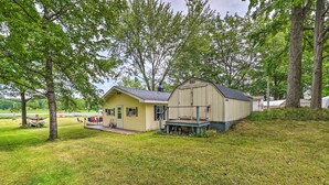 A two-story house with a porch and a garage, surrounded by trees and a grassy yard. - Cottage on Tubbs Lake w/ Fire Pit Near Skiing! (Mecosta)