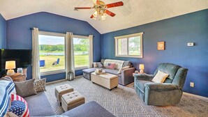 A living room with a ceiling fan, large windows, a sofa, a chair, and a coffee table. - Cottage on Tubbs Lake w/ Fire Pit Near Skiing! (Mecosta)