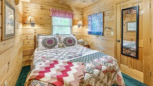 A wooden cabin bedroom with a large bed, two bedside lamps, a window with a view, and a door with a mirror and picture frames. - 11 Mi to Dtwn: Franklin Cabin w/ Porch! (Franklin)