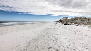 Une vaste plage de sable où l’on peut voir des empreintes de pas menant à l’eau, un bâtiment perché sur une falaise et un ciel partiellement nuageux.