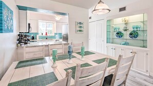 Un coin repas avec une table en bois, des sets de table verts et des chaises blanches. Un meuble vitré contenant des objets décoratifs est accroché au mur.