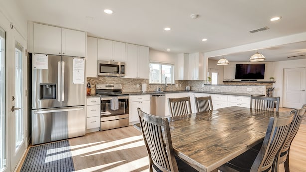 A modern kitchen with stainless steel appliances, a dining area with a wooden table and chairs, and a large TV mounted above the counter. - Private Pool & Patio: Modern Lake Havasu Home! (Lake Havasu City)