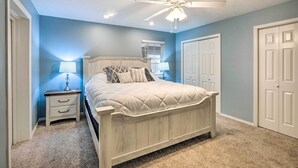 Quarto com cama, mesa de cabeceira, abajur, ventoinha no teto e duas portas.
