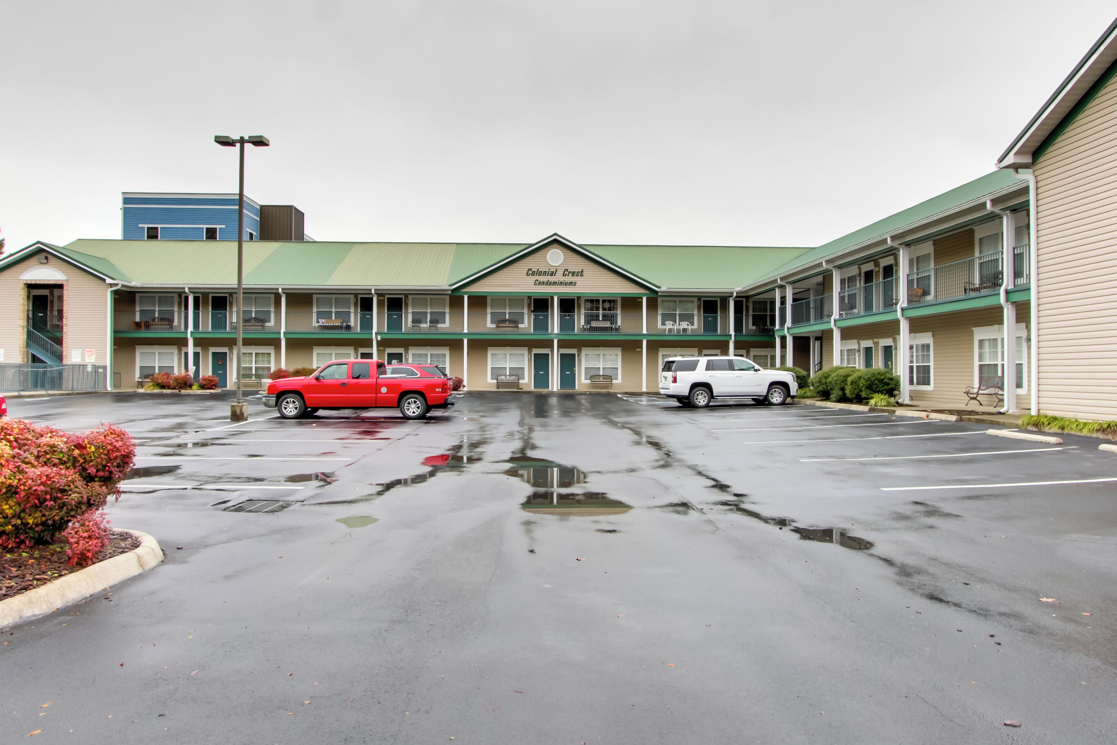 A motel with a green roof and beige exterior, featuring a parking lot with two cars and a sign reading "Colonial Crest Condominiums."