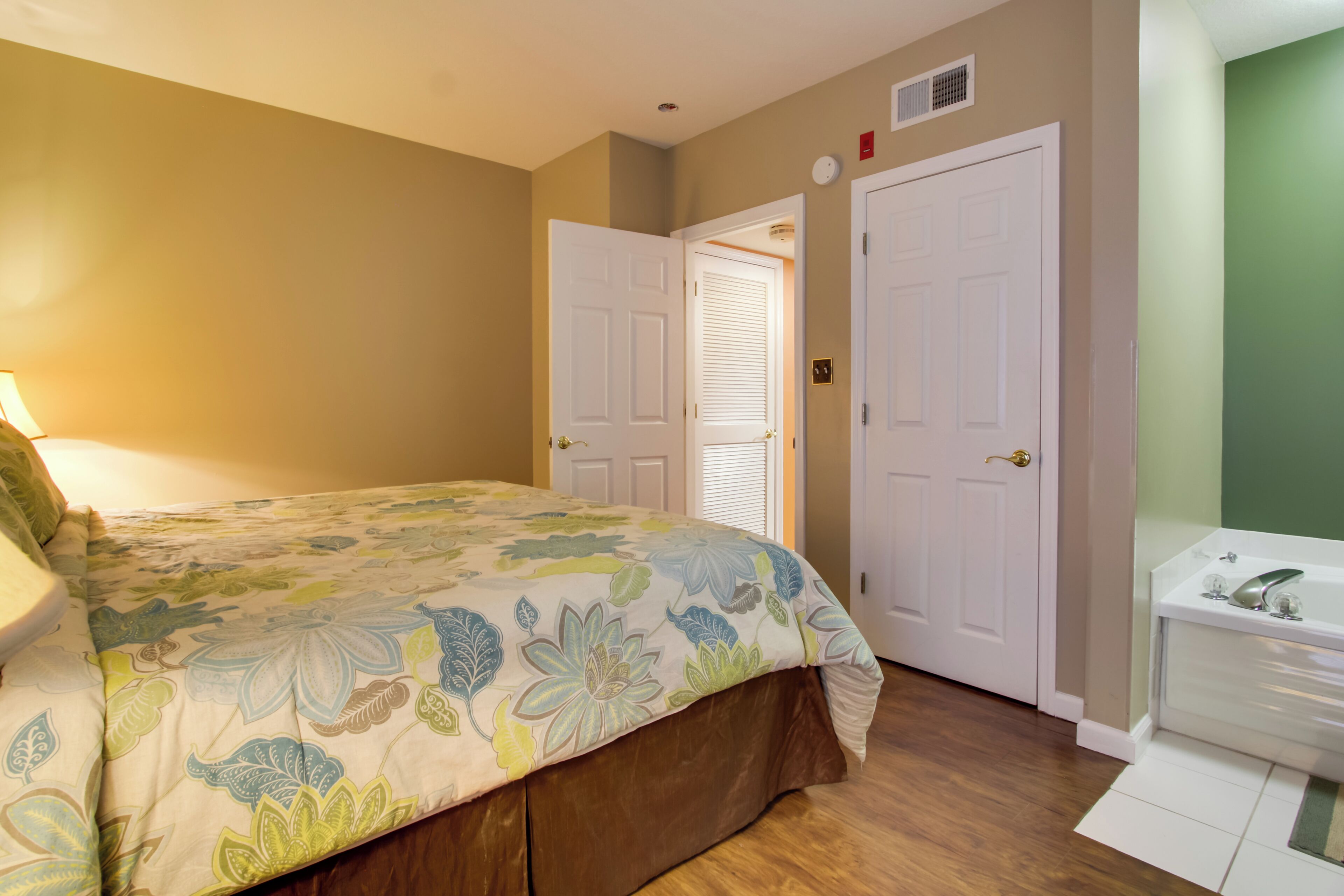 Une chambre à coucher avec une couvre-lit à motifs floraux, une porte blanche et une salle de bain avec un lavabo et un miroir blancs.