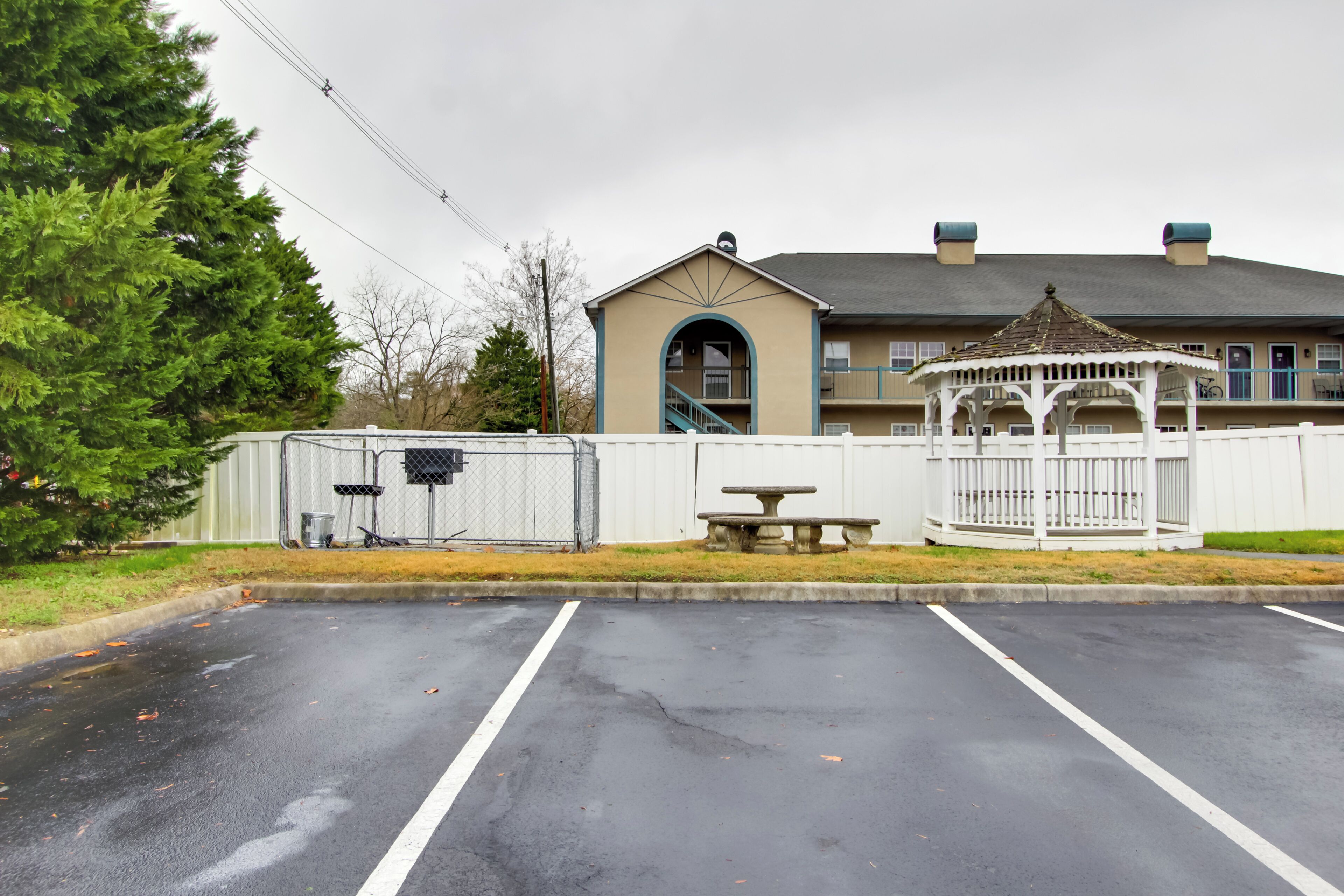 A parking lot with a gazebo and picnic table, a building with a covered entrance, and a fenced area with a trash can.