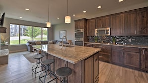 A modern kitchen with dark wood cabinets, a large island with bar stools, stainless steel appliances, and a view of a residential area through large windows. - Modern Huntsville Resort Retreat w/ Deck & Hot Tub (Huntsville)