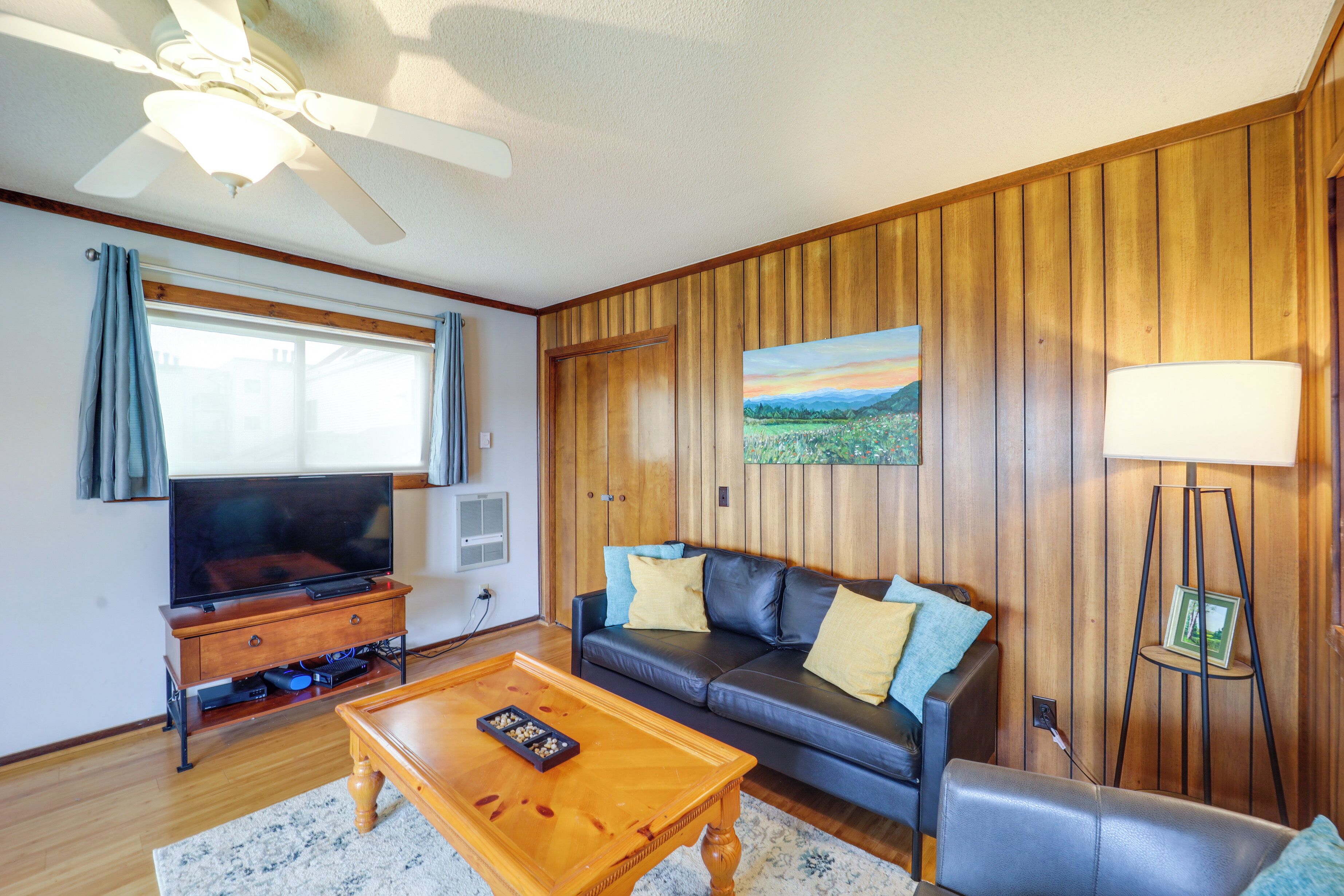 Ein Wohnzimmer mit einer holzverkleideten Wand, einem schwarzen Ledersofa, einem Couchtisch aus Holz, einem Fernseher auf einem Holzständer und einem Deckenventilator.