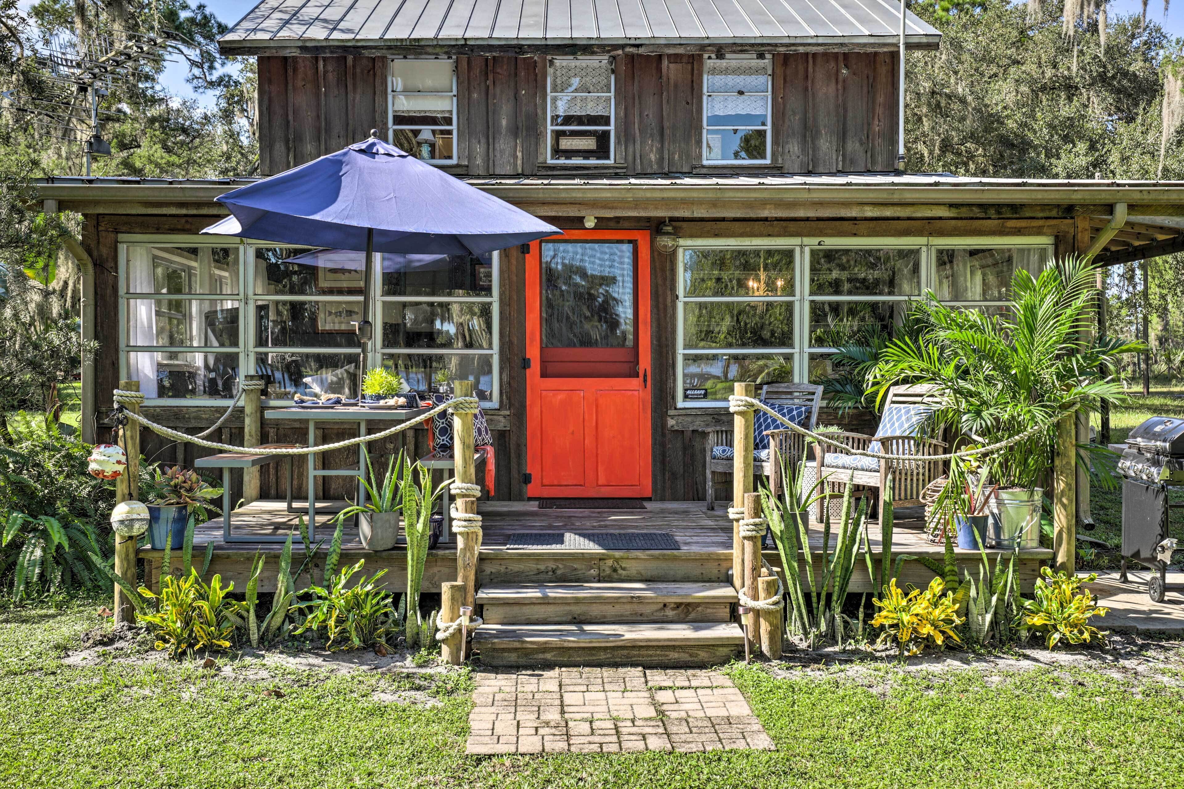 A rustic wooden house with a red door, a covered patio with a blue umbrella, and a garden with various plants.