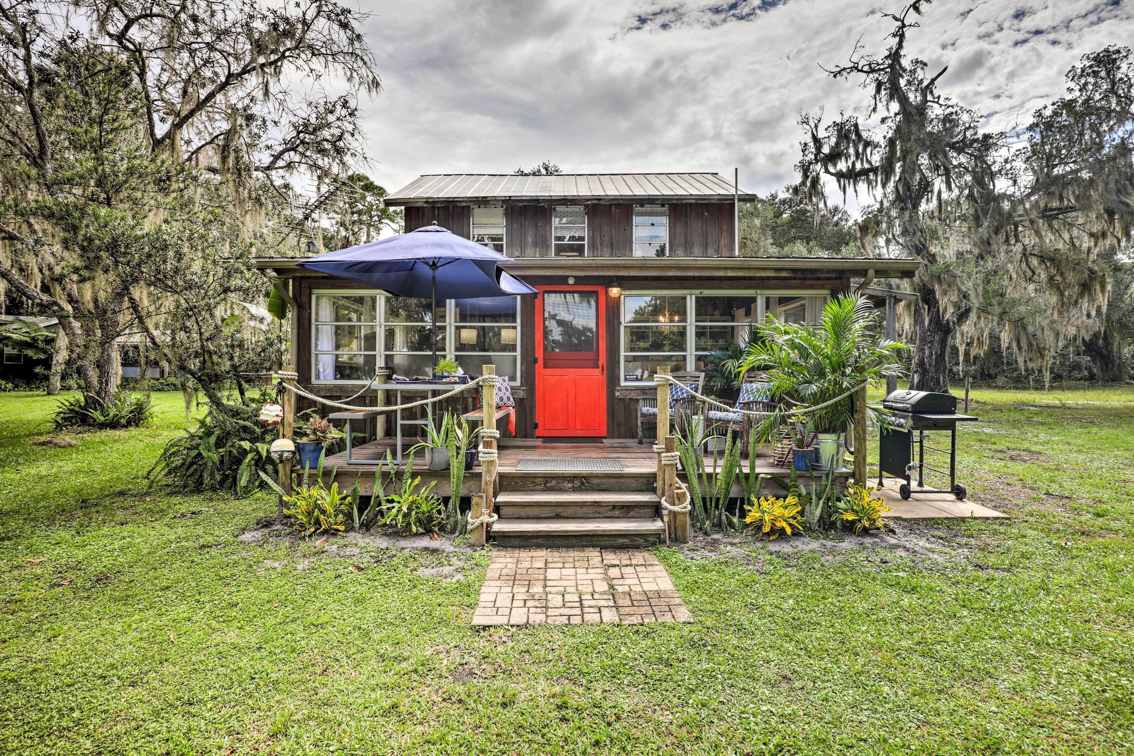 A rustic wooden building with a red door, surrounded by a well-maintained lawn and various plants.