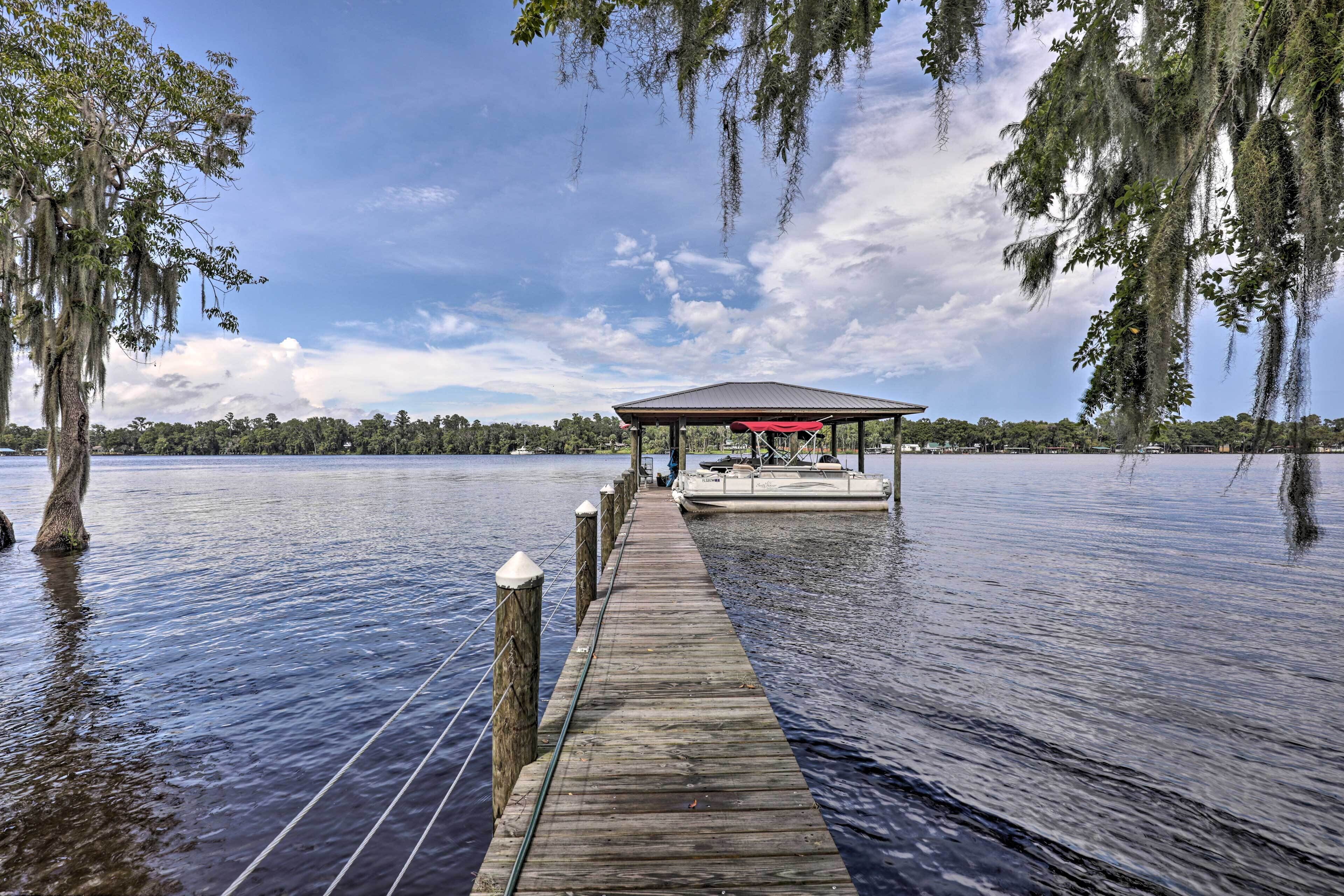 Une jetée en bois mène à un pavillon abrité situé au bord d’un lac paisible, entouré d’arbres et surplombé d’un ciel dégagé.