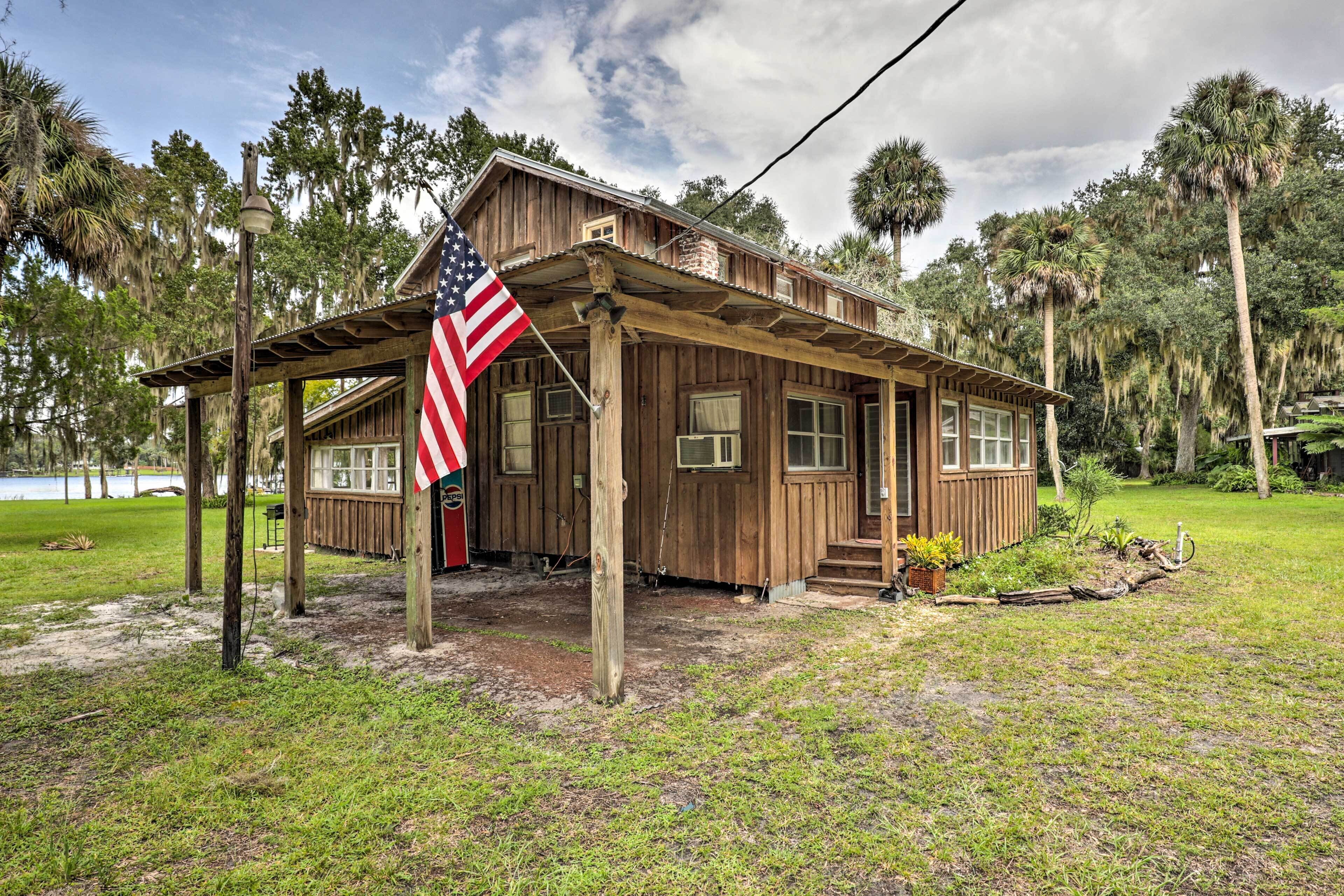 A wooden cabin with a porch, an American flag, and a view of a lake and trees.