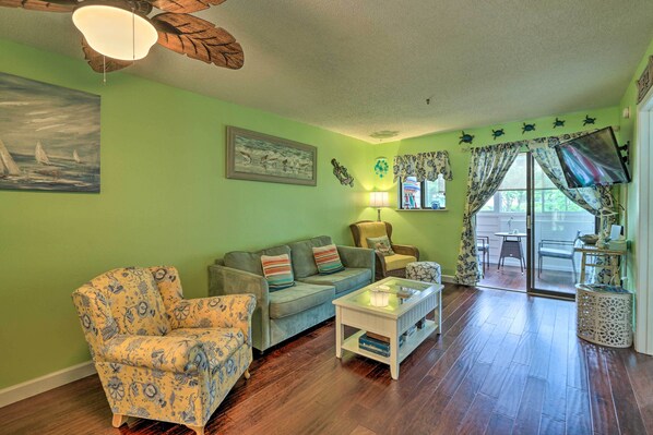 A living room with a green wall, wooden flooring, a sofa, a chair, a coffee table, a lamp, and a TV. - Beach + Pool Access: Colorful Hilton Head Condo! (Hilton Head Island)