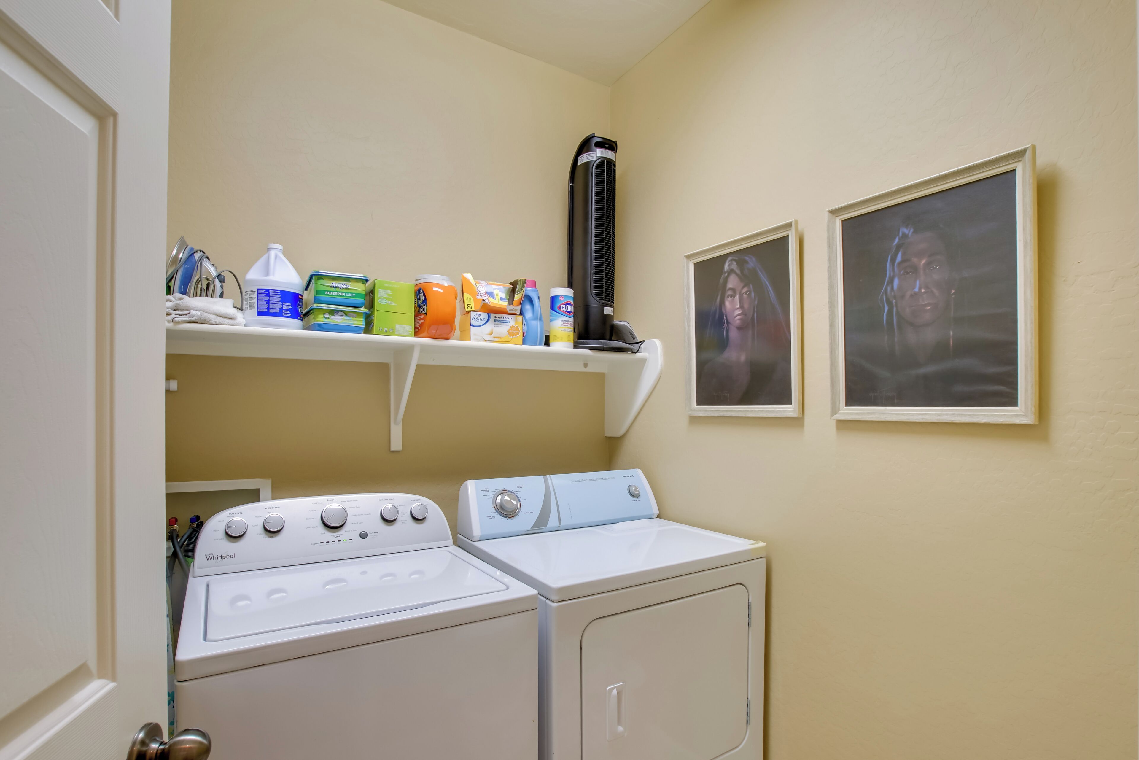 A laundry area with a washer and dryer, a shelf with cleaning supplies, and two framed pictures on the wall.
