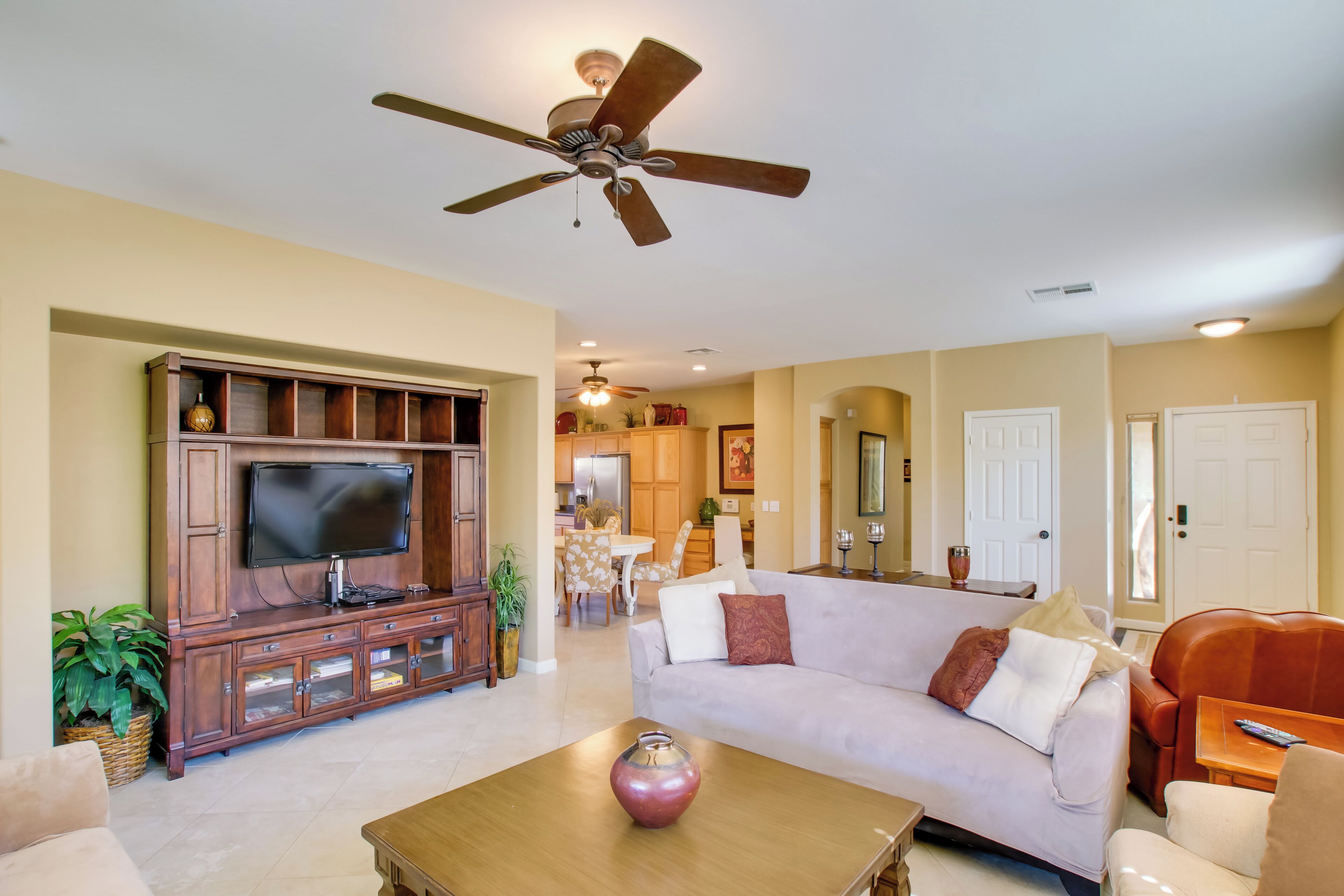 A living room with a flat-screen TV, wooden entertainment center, a ceiling fan, a sofa, a coffee table, and a dining area in the background.