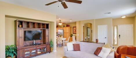 A living room with a flat-screen TV, wooden entertainment center, a ceiling fan, a sofa, a coffee table, and a dining area in the background.