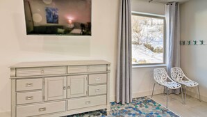 A room with a white dresser, a flat-screen TV mounted on the wall, two white chairs, a window with a view of trees, and a patterned rug. - Anchorage Studio Escape ~ 14 Miles From Downtown! (Anchorage)
