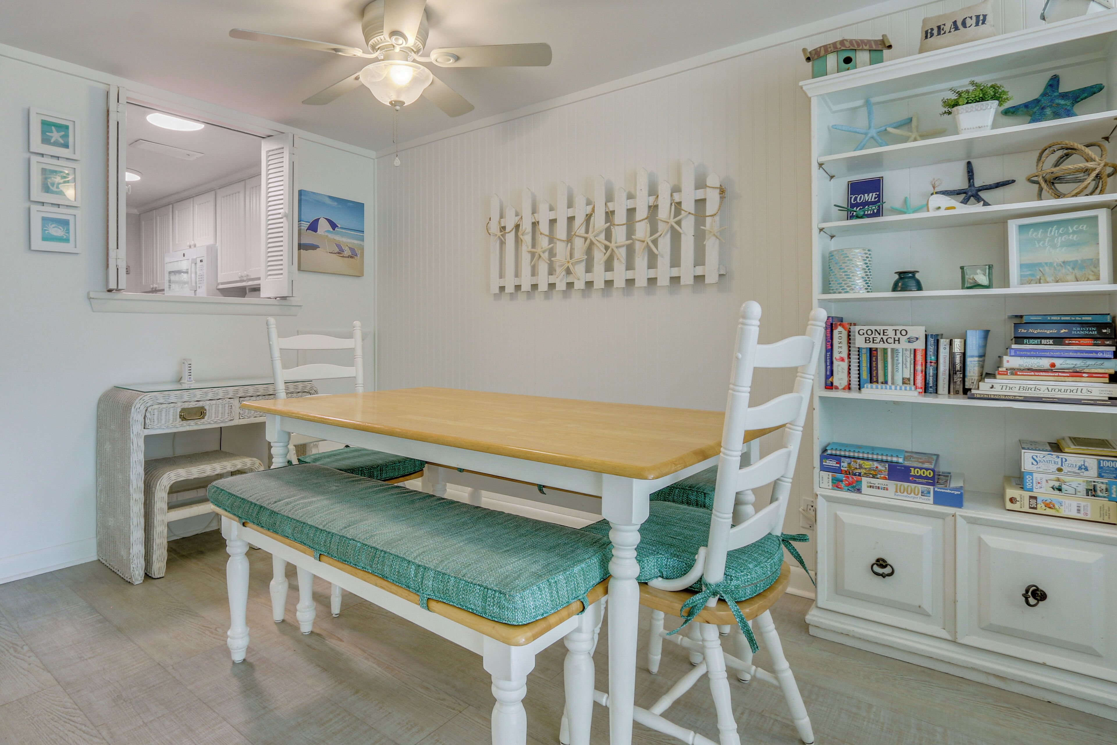 A dining area with a wooden table and chairs, a bookshelf with books and decorative items, and a white picket fence wall art.