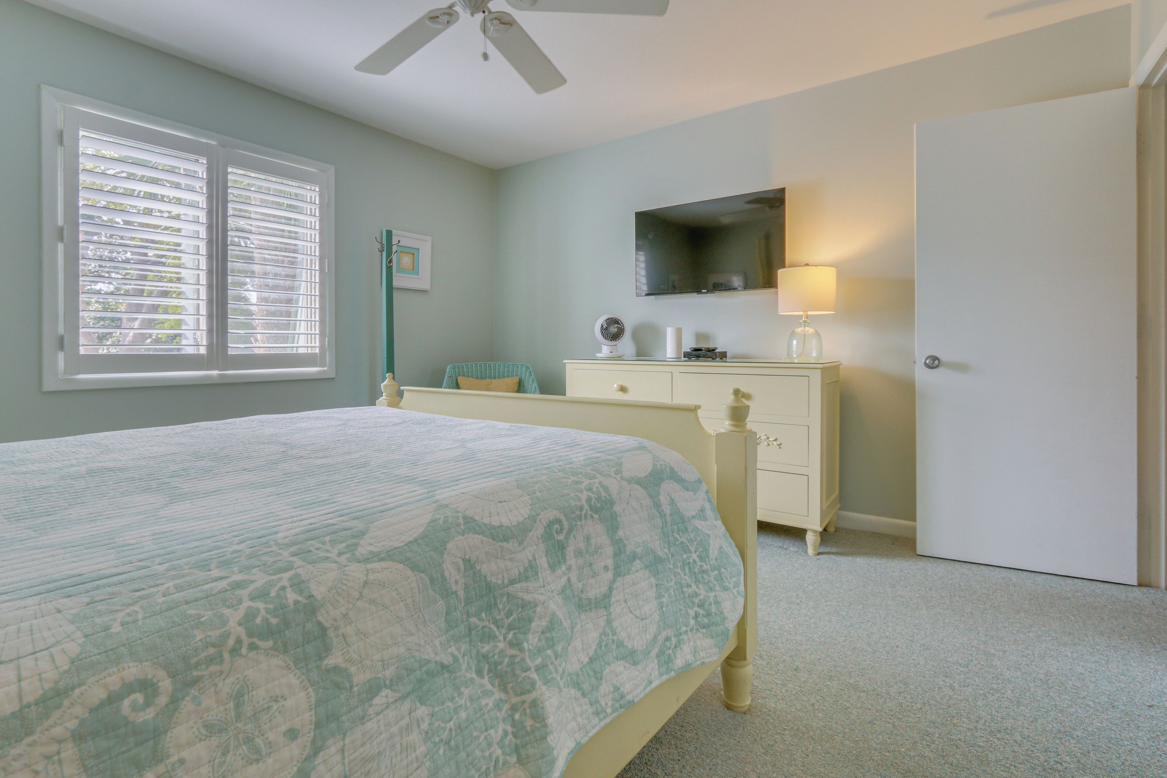 Une chambre à coucher comprenant un lit, une commode, un miroir, un ventilateur de plafond et une fenêtre avec des stores.
