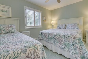 Une chambre avec deux lits, une fenêtre avec des stores, une table de chevet avec une lampe et un ventilateur de plafond.