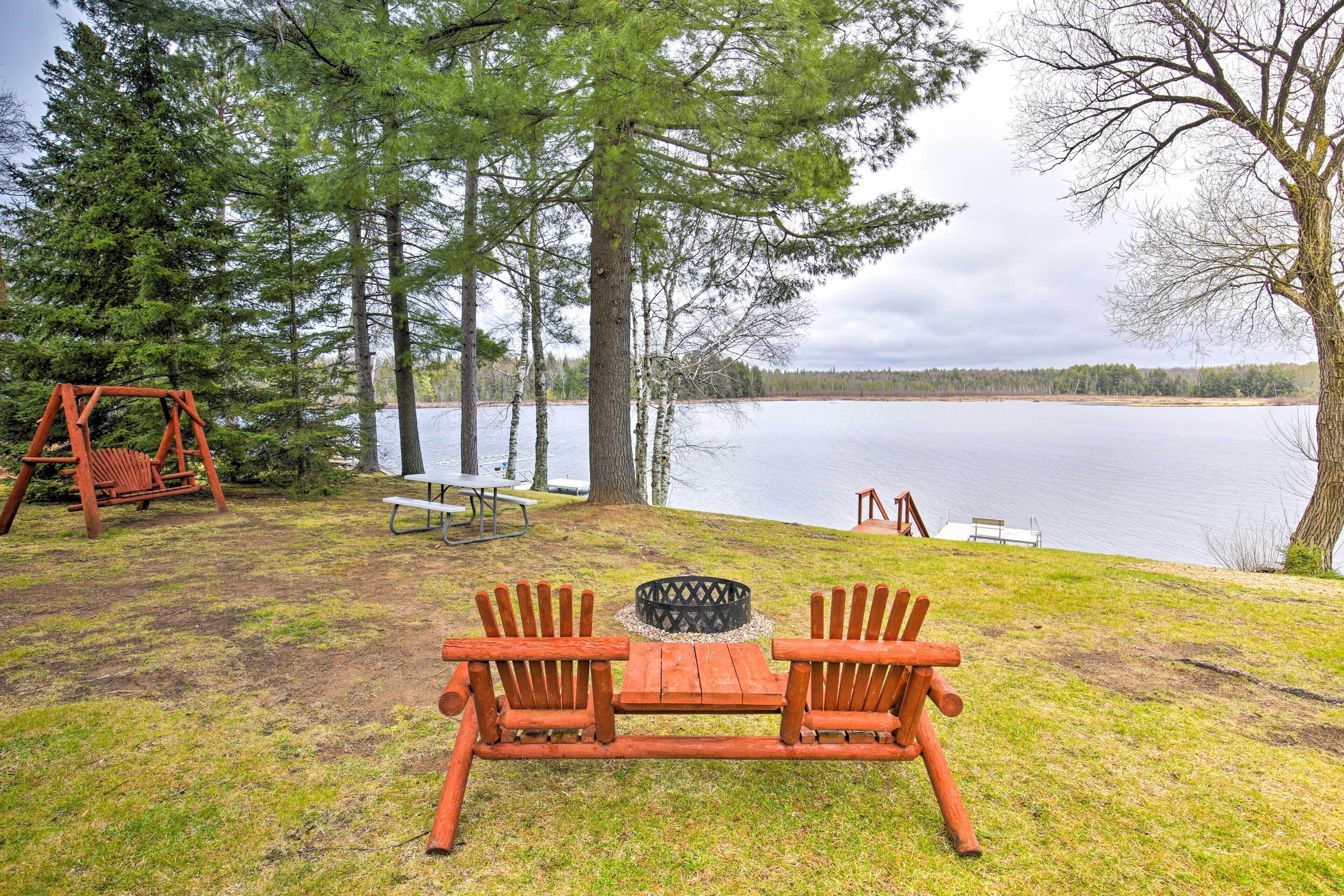 Un espacio al aire libre junto al lago con mesa y sillas de madera, un conjunto de columpios y una mesa de picnic.