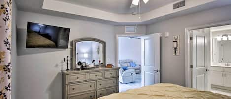 A bedroom with a bed, dresser, mirror, ceiling fan, and a door leading to another room.
