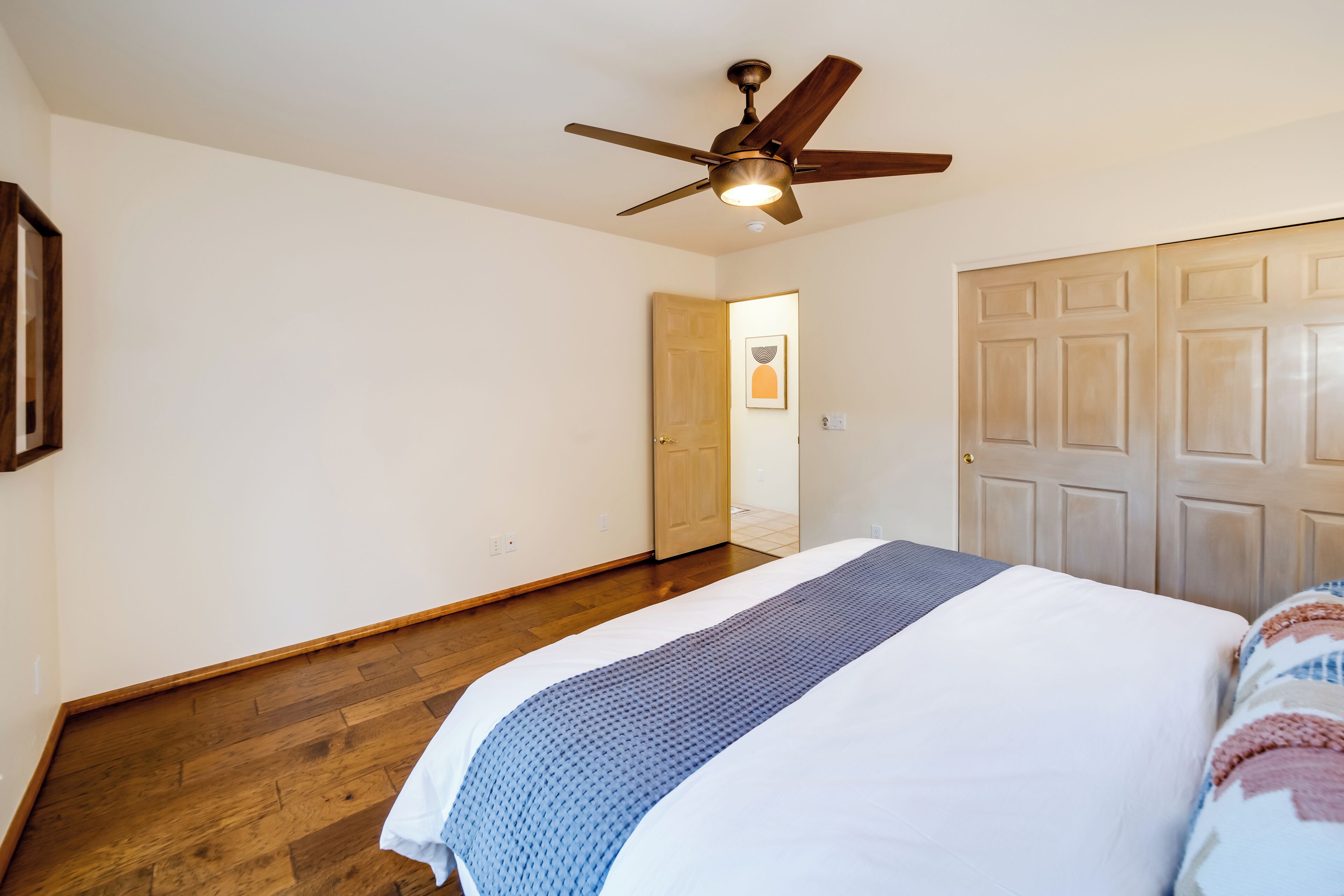 A bedroom with a wooden floor, a bed with a blue and white bedspread, a ceiling fan, and a door leading to another room.