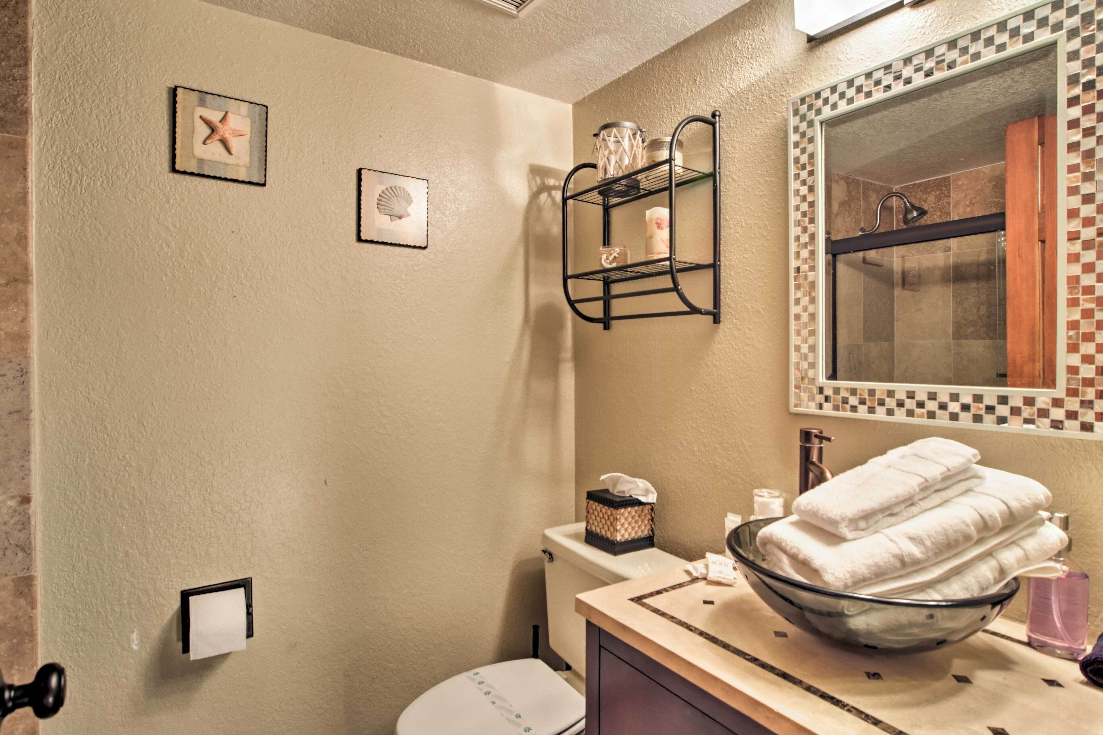 A bathroom with a toilet, sink, and a glass bowl filled with towels. There are framed pictures on the wall and a shelf with toiletries.