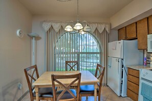 Un coin repas avec une table et des chaises en bois, un lustre, un réfrigérateur et une fenêtre avec des rideaux.
