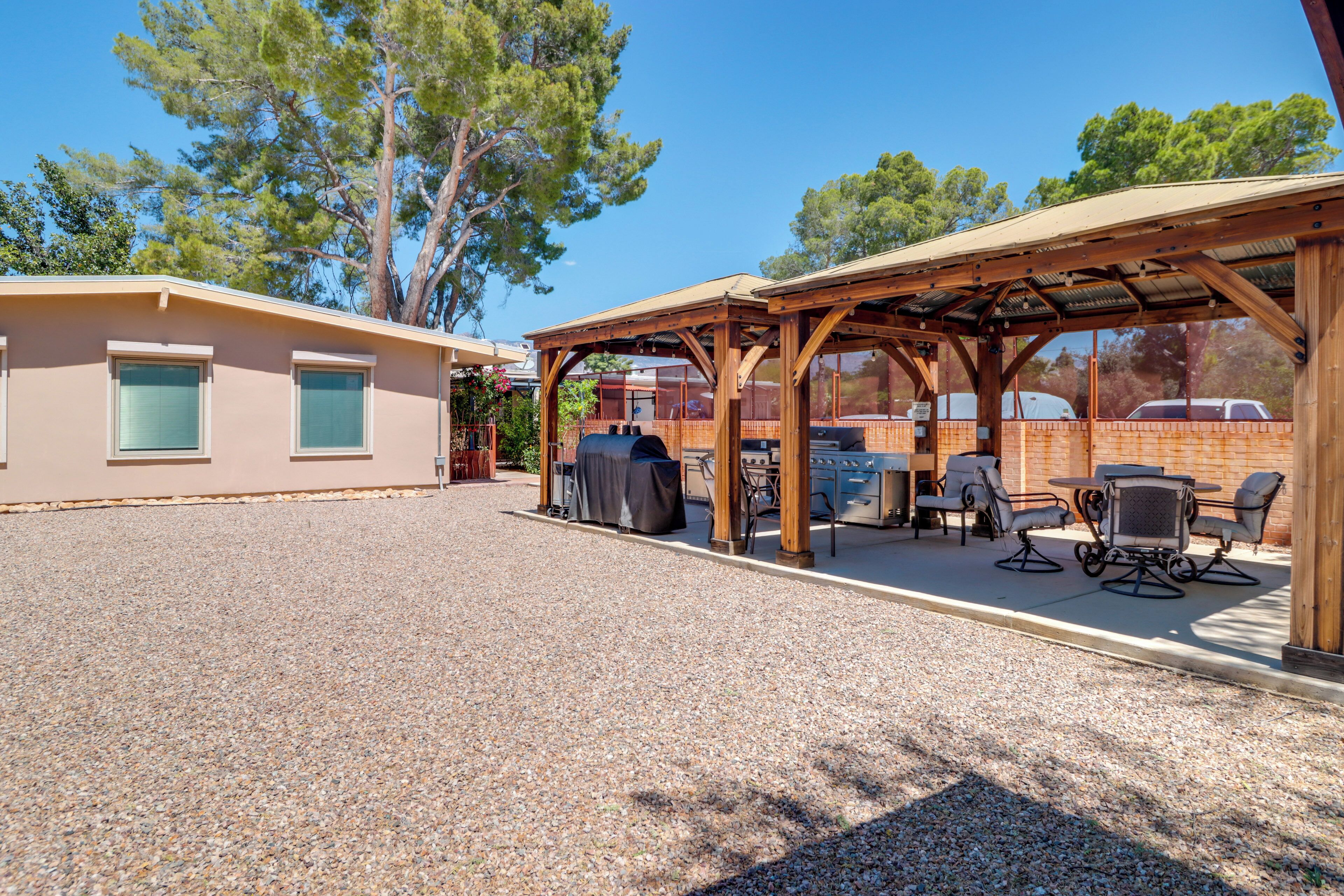 A covered outdoor seating area with tables and chairs, a barbecue grill, and a building with windows.