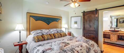 A bedroom with a large bed, a ceiling fan, a wooden door, and a bathroom with a sink and mirror.