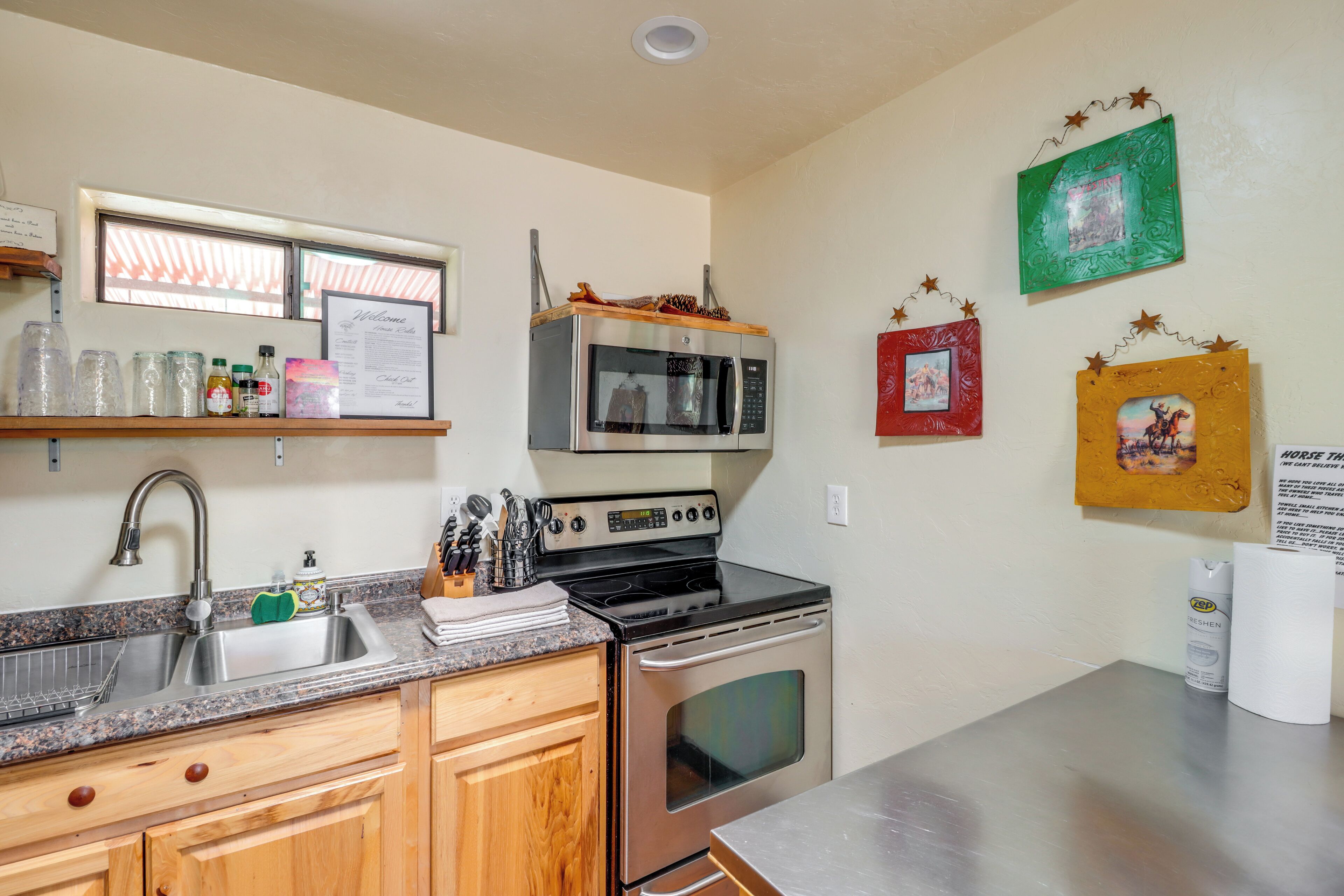 A kitchen with a stainless steel sink, a microwave, an oven, wooden cabinets, and a window with blinds.