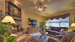 A living room with a stone fireplace, a ceiling fan, a large flat-screen TV, a sofa, a coffee table, and a large window with blinds. - Arizona Home w/ Patio, Fire Pit & Gas Grill (Williams)