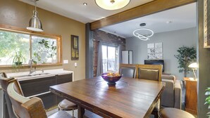Un comedor con una mesa de madera, sillas, un bol de fruta, una ventana con plantas y una cocina al fondo.