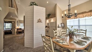 Un coin repas avec une table en bois dressée pour quatre personnes, entourée de chaises blanches. Un lustre est suspendu au-dessus de la table, et une fenêtre avec des rideaux est visible à l’arrière-plan.