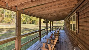 Un espacio de estar al aire libre cubierto con bancos y mesas de madera, baranda de madera y vista a un campo cubierto de césped y árboles.