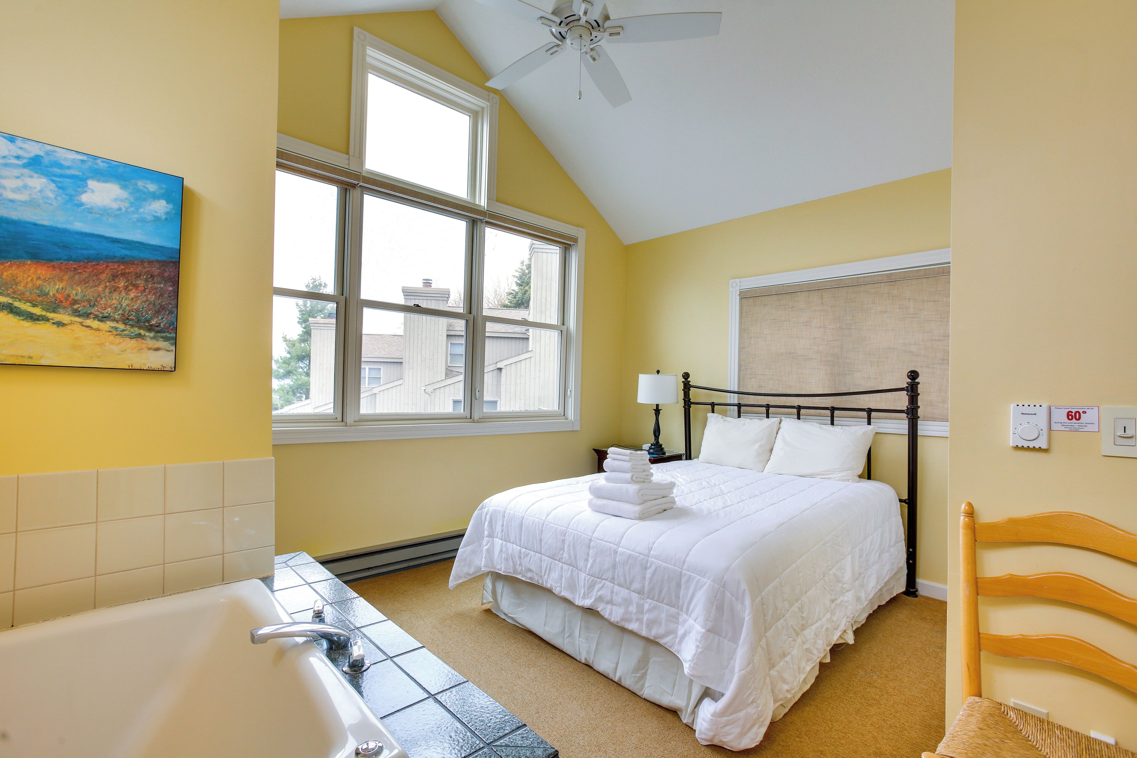 A bedroom with a bed, a window, a ceiling fan, a bedside table, and a wall-mounted thermostat.