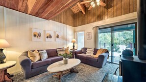 A cozy living room with a wooden ceiling, a fireplace, and a large window offering a view of the outdoors.